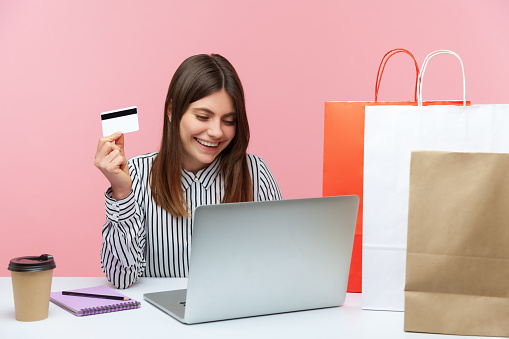 Happy excited woman sitting at laptop and holding credit card, doing shopping online, paper bags lying on desk, fast delivery service. Indoor studio shot isolated on pink background