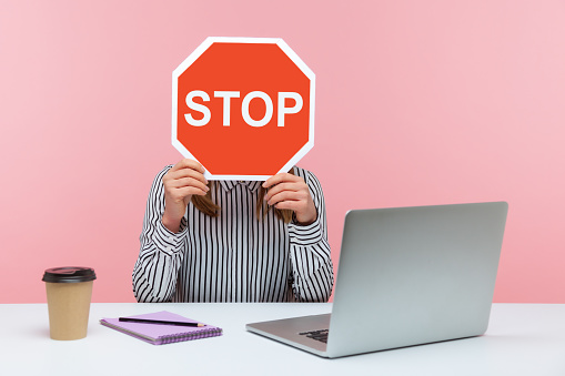 Female office worker in striped shirt sitting at workplace hiding face behind red stop traffic sign avoiding conflicts, afraid of workplace bullying. Indoor studio shot isolated on pink background
