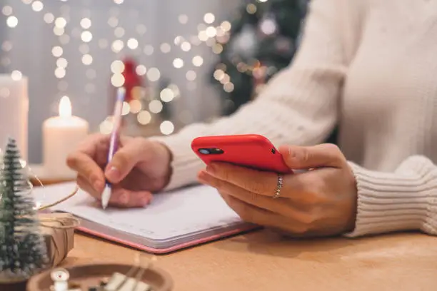 Photo of Woman hands with pen and mobile phone writing christmas wish list, goals.