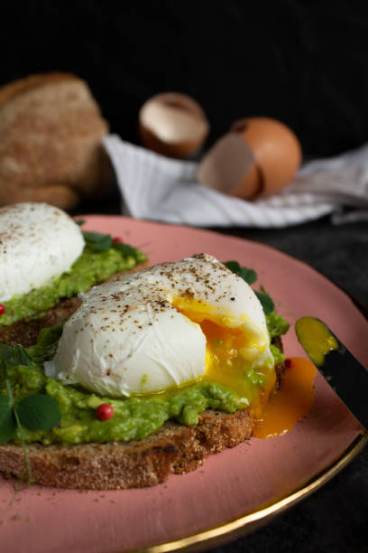 istock Poached egg cut with a knife on avocado puree on toast bread on a pink plate. Egg scarlop, bread and towel on background 1287587099