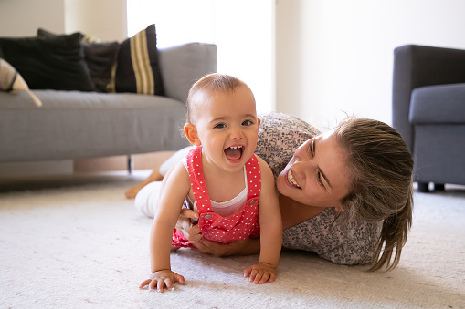 Joyful mom and little girl lying on carpet in living room. Happy blonde mom playing with cheerful daughter at home and smiling. Cute baby laughing with open mouth. Motherhood and weekend concept