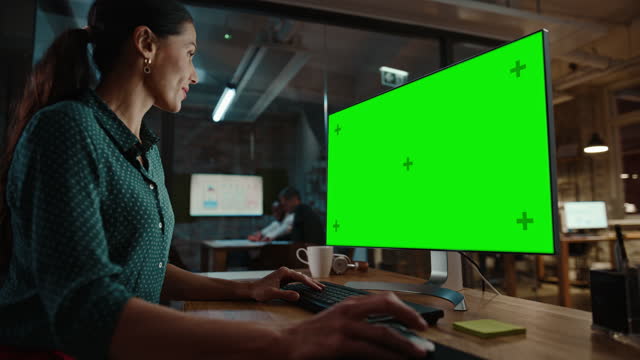 Middle Aged Multiethnic Specialist Working on Desktop Computer with Green Screen Mock Up Display in Busy Creative Office. Beautiful Diverse Female Manager in Green Polka Dot Blouse.