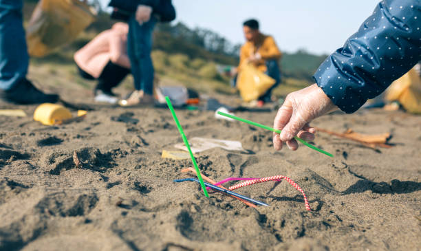 Woman picking up straws on the beach Woman hand picking up straws on the beach with group of volunteers working in the background. Selective focus in straws in foreground straw stock pictures, royalty-free photos & images