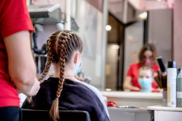 Woman making braid to Teenage Girl sitting in front of the mirror. Females wearing face mask during Corona virus pandemic