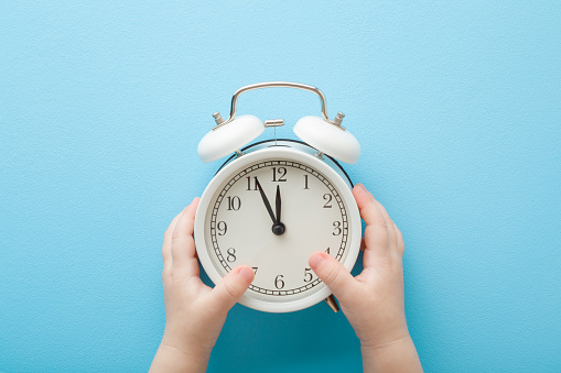 Baby hands holding white alarm clock on light blue table background. Pastel color. Time concept. Closeup. Point of view shot. Top down view.