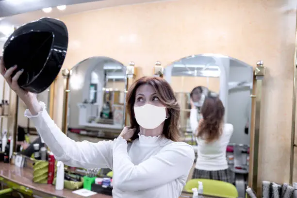 Woman using Mirror looking at new hairstyle in Hairdresser. Long Hair Female with face mask during Coronavirus Pandemic in Beauty Salon
