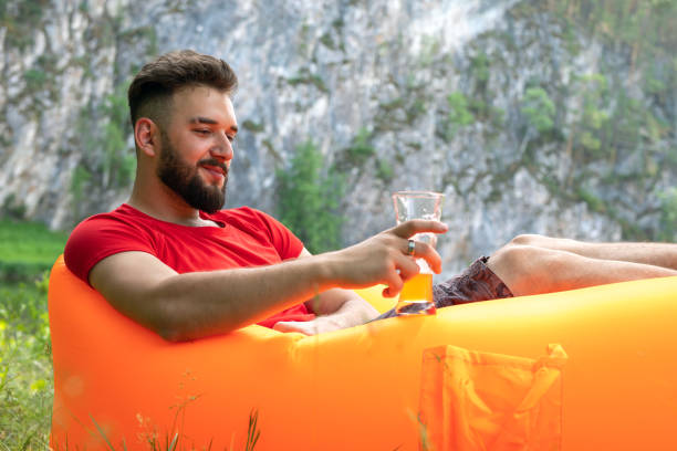istock Side view of handsome young man drinking beer while sitting at inflatable air lounger in park 1287601301