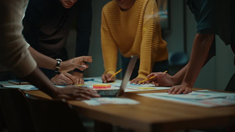 Close Up of Diverse Multiethnic Team Having Conversation in Meeting Room in a Creative Office. Colleagues Lean On a Conference Table, Look at Laptop Computer and Make Notes with Pencils on Notebooks.