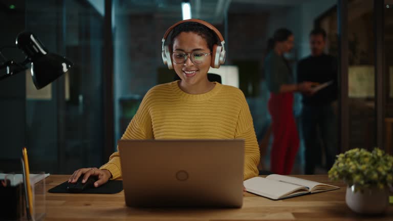 Young Latin Project Manager Putting On Headphones and Working on Laptop Computer in Busy Creative Office Environment. Beautiful Diverse Multiethnic Female Specialist is Writing Business Strategy.