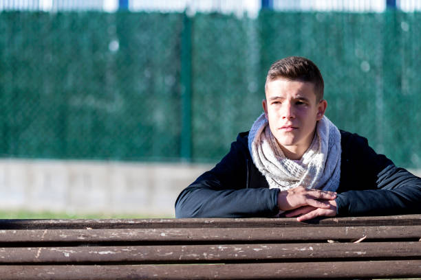 Attractive young guy in city park, leaning on wood bench while looking away