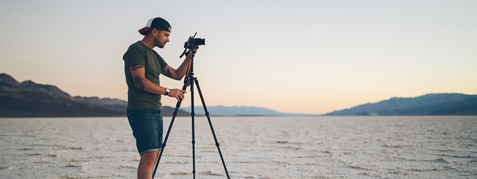 Side view of full body male traveler in cap standing near tripod and photographing  sunset over picturesque highland while resting in Death Valley