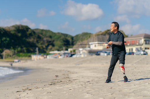 A senior adult man with an artificial leg is fishing at the beach.