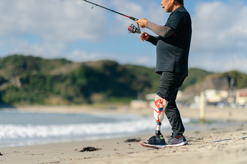 A senior adult man with an artificial leg is fishing at the beach.