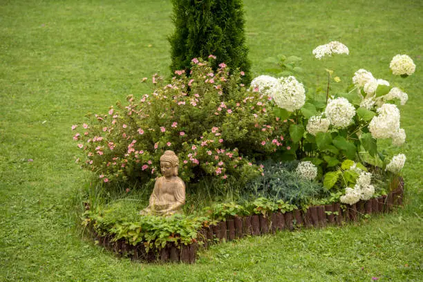 Photo of Meditating Buddha figure sit inside small island of flowers in flower bed, surrounded by natural wood roll palisade.