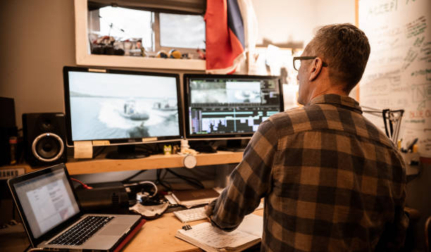 Mature man working at his home editing suite during the pandemic lock-down.. He is editing video content on professional editing computer system.