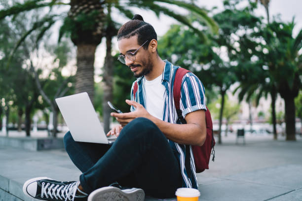 istock Positive young black hipster male using laptop and smartphone while sitting on step in street 1287619636
