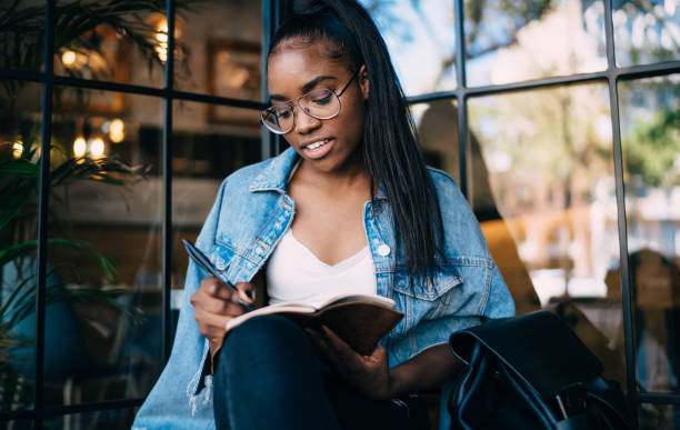 istock Clever beautiful dark skinned student in eyewear drawing in notepad enjoying spending time on hobby, positive dark skinned female student making notes for learning in cafe interior with notebook 1287622743