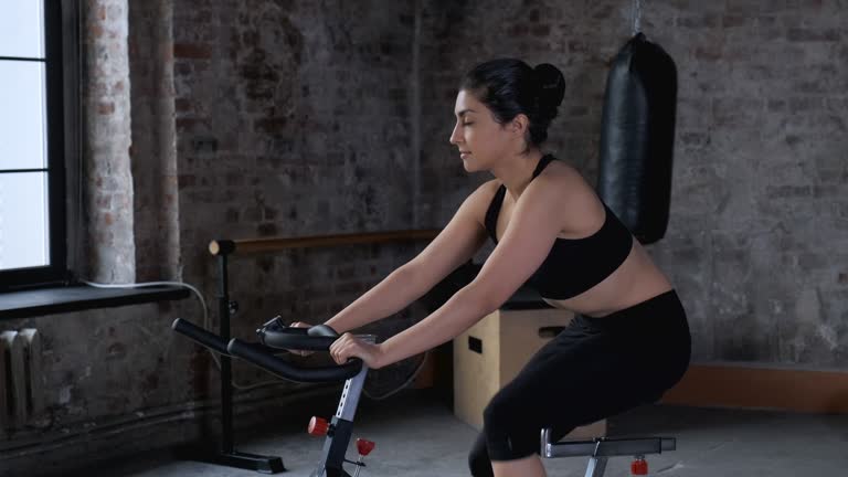 Young Athletic Indian Woman Doing a Workout, Engaged in Cycling Dressed in Sportswear Black Top and Leggings, is In the Gym