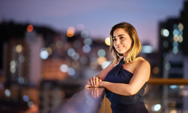 jeune femme de sourire se penchant sur une balustrade de balcon au crépuscule - evening wear photos et images de collection