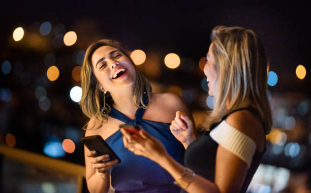 amis élégants restant sur un balcon la nuit et riant d’un message - tenue de soirée photos et images de collection