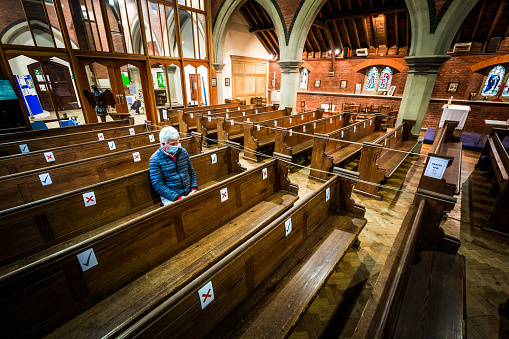 Color image depicting a senior man in his 60s sitting alone in a church while wearing a protective face mask and face shield. The pews are mark with ticks and crosses to delineate a social distancing system during the covid-19 pandemic. Room for copy space.