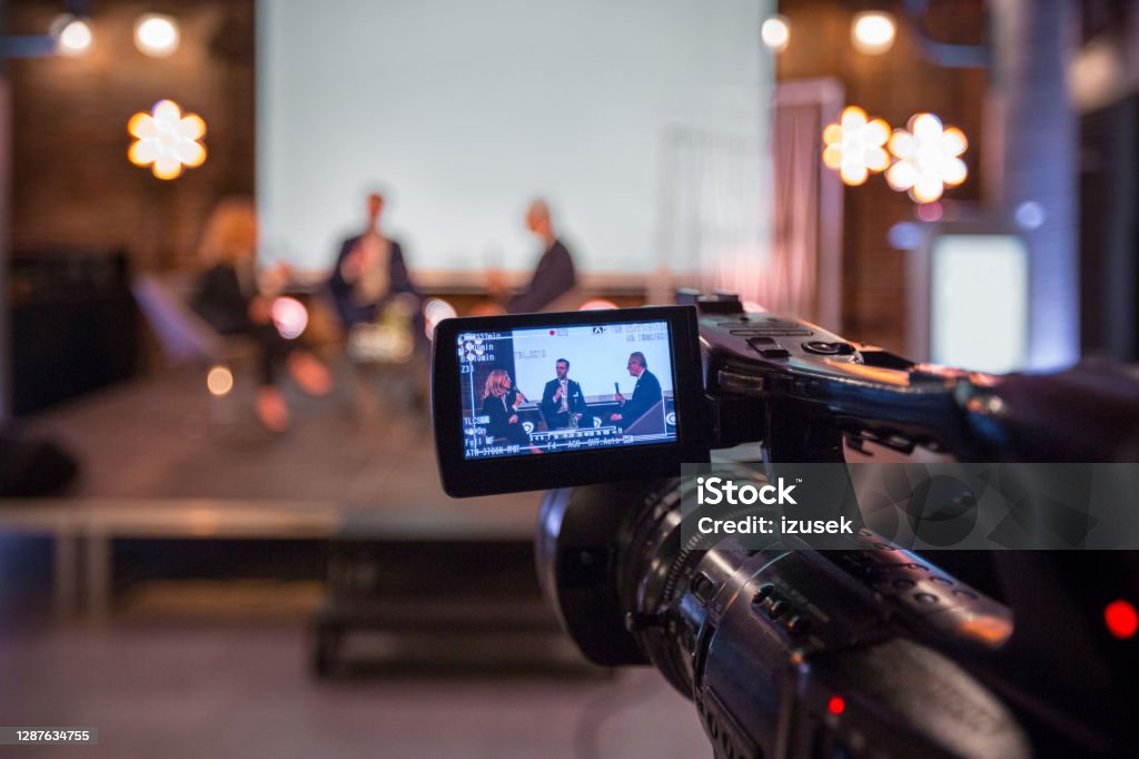 Businesswoman and businessmen during online seminar Businesswoman and businessmen discussing during online seminar, sitting on armchairs on the stage. Focus on video camera. Home Video Camera Stock Photo