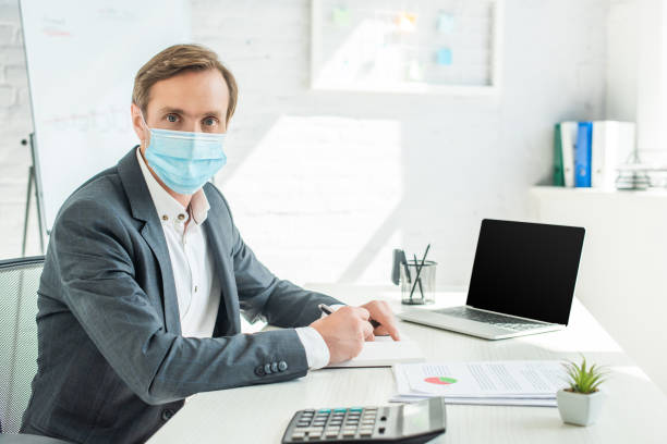 istock Businessman in medical mask looking at camera, while writing in notebook at workplace on blurred background 1287637363