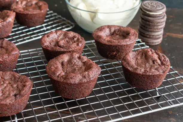 Brownie cups filled with a sandwich cookie wire cooling rack with a bowl of frosting in the background