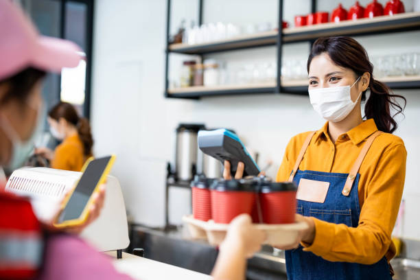 mobile payment concept Asian young woman clerk wearing face mask delivers coffee through delivery staff - pays by contactless phone to credit card system asian cashier stock pictures, royalty-free photos & images