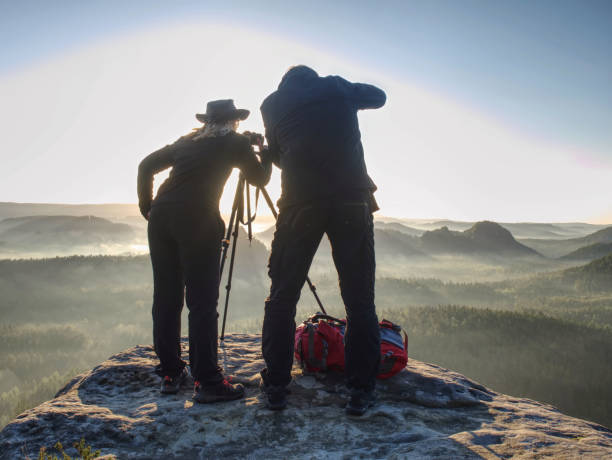 山の頂上で写真カメラを持つカップルの観光客は、自然の中でハイキング中に屋外で夕日を見る - climbing mountain climbing rock climbing women ストックフォトと画像
