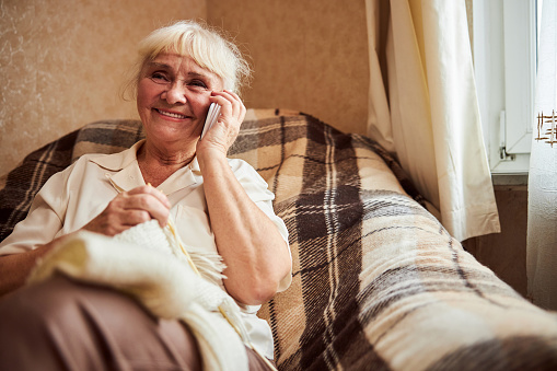 Joyful elderly lady having phone conversation and smiling while sitting in armchair and knitting