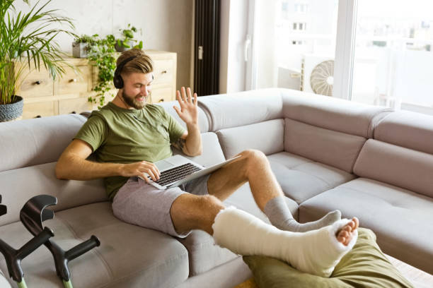 Cheerful young man with broken leg during video call Smiling young man with broken leg in plaster cast lying down on sofa at home, using laptop and having video conference. broken leg stock pictures, royalty-free photos & images