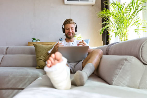 Cheerful young man with broken leg during video call Smiling young man with broken leg in plaster cast lying down on sofa at home, using laptop and having video conference. broken leg stock pictures, royalty-free photos & images