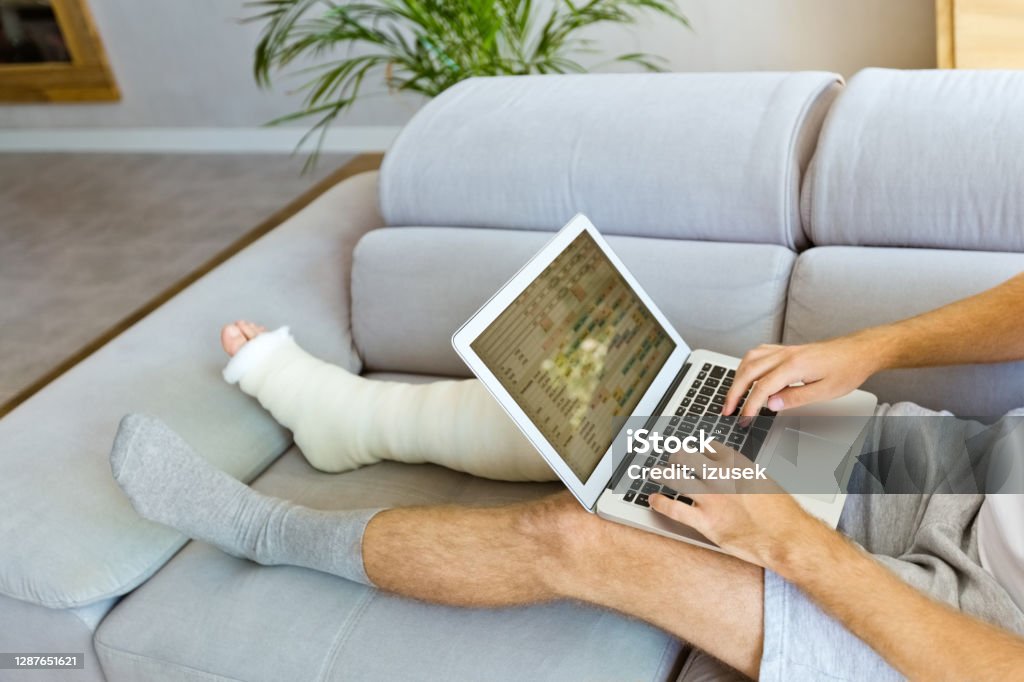 Man with broken leg using laptop Man with broken leg in plaster cast lying down on sofa at home, using laptop. Unrecognizable person. Orthopedic Cast Stock Photo