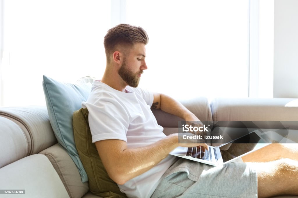 Young man using laptop at home Young man lying down on sofa at home, using laptop, working at home. 20-24 Years Stock Photo