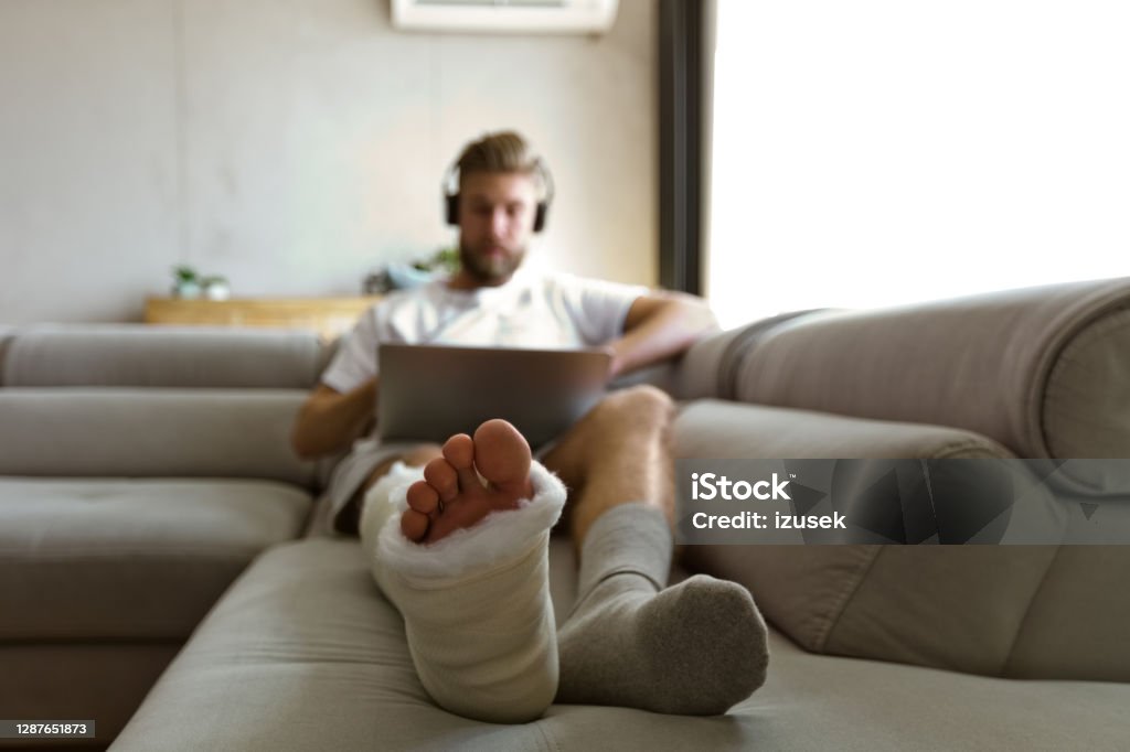 Young man with broken leg during video call Young man with broken leg in plaster cast lying down on sofa at home, using laptop and having video conference. Focus on leg. Orthopedic Cast Stock Photo