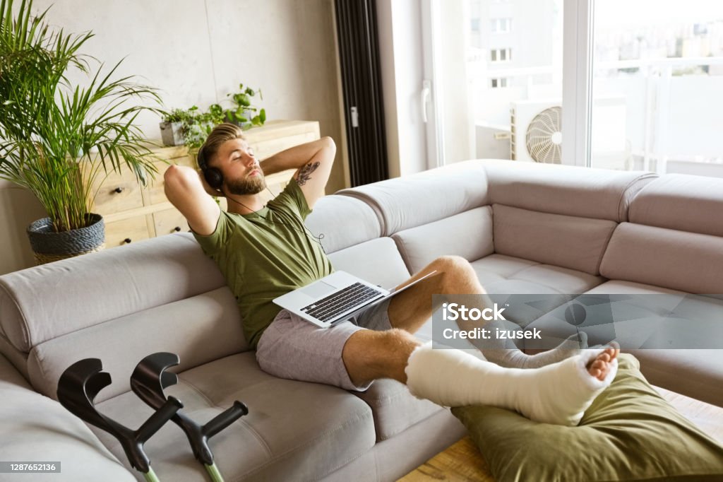 Young man with broken leg resting at home Young man with broken leg in plaster cast lying down on sofa at home, using laptop. High angle view. Orthopedic Cast Stock Photo
