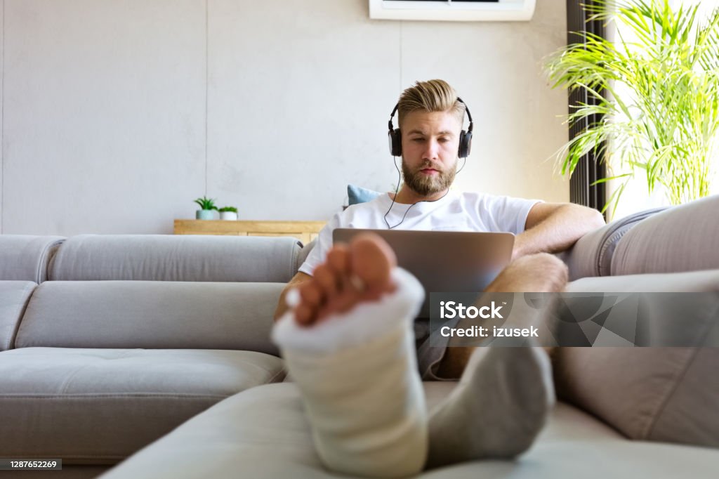Young man with broken leg using laptop Young man with broken leg in plaster cast lying down on sofa at home, using laptop and having video conference. Misfortune Stock Photo