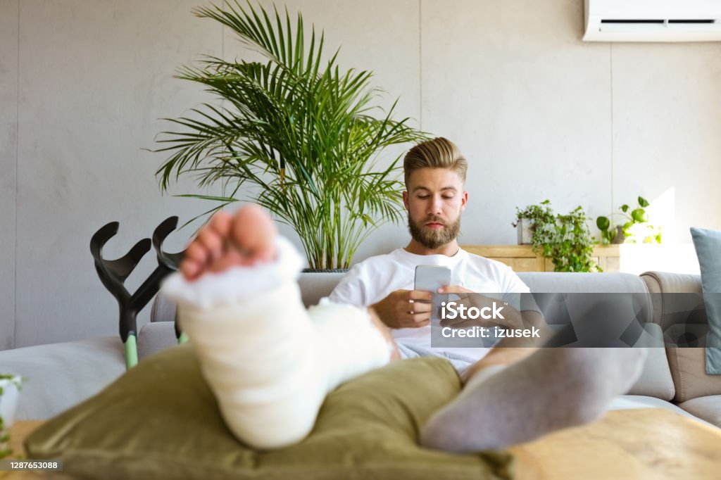Young man with broken leg using smart phone Young man with broken leg in plaster cast lying down on sofa at home, using smart phone. Broken Leg Stock Photo