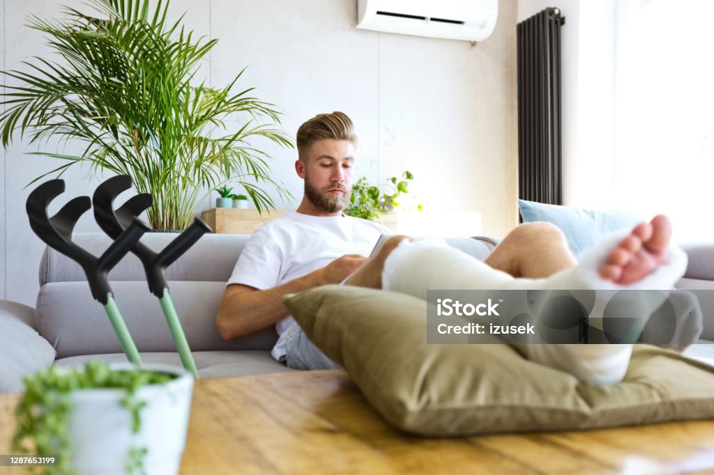 Young man with broken leg using smart phone Young man with broken leg in plaster cast lying down on sofa at home, using smart phone. Broken Leg Stock Photo