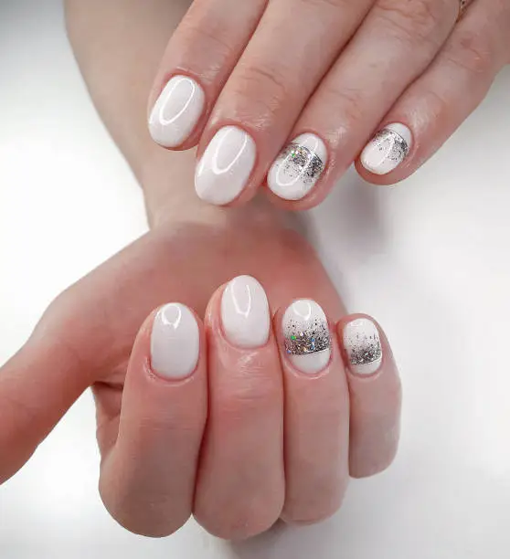 White nails with sequins and a silver crown design on a white background. White shiny gel polish on round short nails