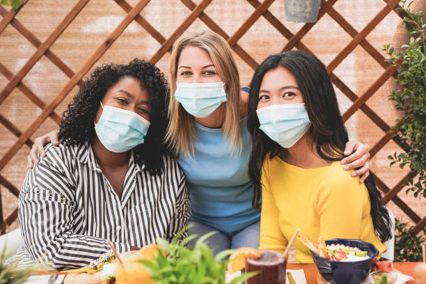 istock Young multiracial girls looking in camera while wearing surgical face mask for coronavirus - Women enjoy brunch at bar 1287657380