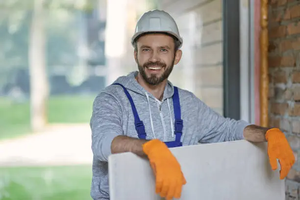 Portrait of handsome young male builder in hard hat looking positive, holding drywall while working at construction site. Building house, profession, safety concept