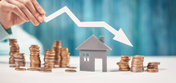 mujer sostiene una flecha hacia abajo sobre el modelo de la casa y la pila de monedas. - vivienda precio fotografías e imágenes de stock