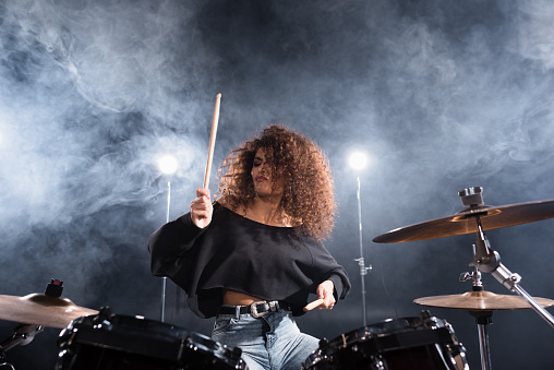 Curly woman with drumsticks playing on drum kit with smoke and backlit on background