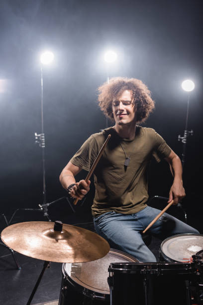 istock Curly musician playing while sitting at drum kit with drumsticks with back light on background 1287685462