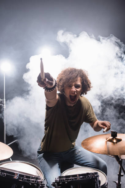 istock Curly drummer pointing with drumstick while shouting and sitting at drum kit with smoke on background 1287685463