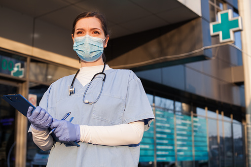 Joven doctora de pie frente al centro de salud, usando mascarilla protectora y equipo de EPI, sosteniendo portapapeles de pacientes médicos, crisis de pandemia COVID-19 photo