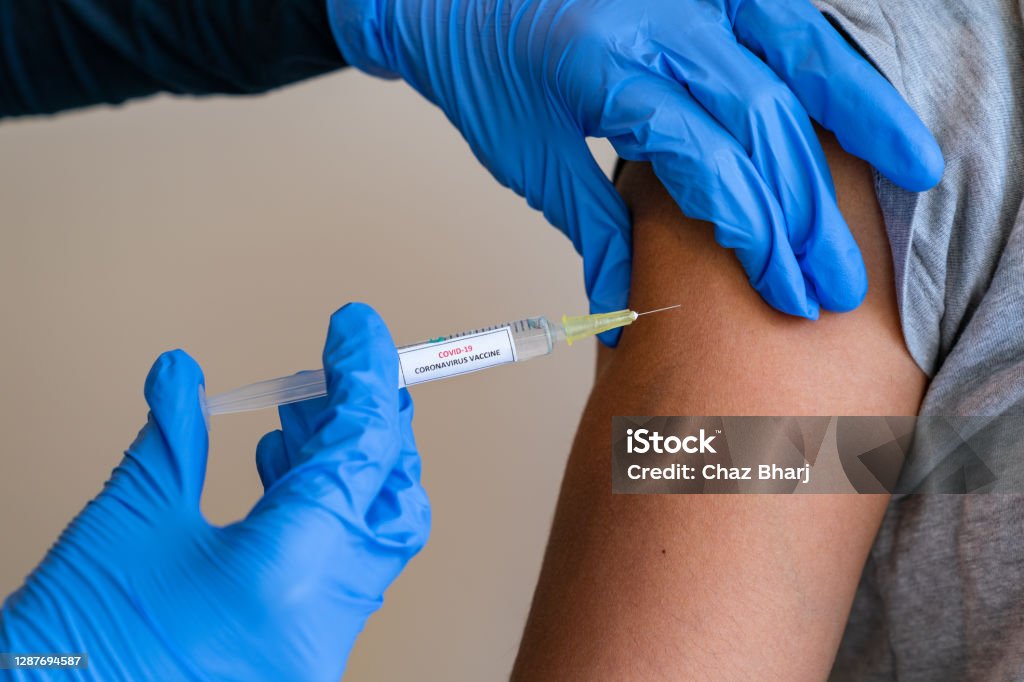 Female doctor wearing blue latex gloves injecting a child in her arm with a needle and syringe containing a dose of the COVID-19 vaccine cure by way of immunisation - 052 This picture depicts a young female clinician using a syringe to inject a concept COVD-19 liquid vaccine into a young girl patient during the Phase 3 vaccination human trials. Vaccination Stock Photo