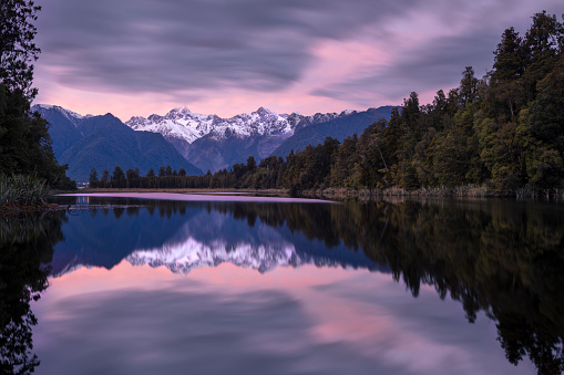 Lake Matheson, New Zealand, is known for it's clear reflections of the Southern Alps, in particular of Mount Tasman (left) and Mount Cook New Zealand's tallest mountain (right). In this photo, the sky is filled with colour before sunrise and the scene is reflected in the still waters of the lake. This lake is a very popular tourist destination.